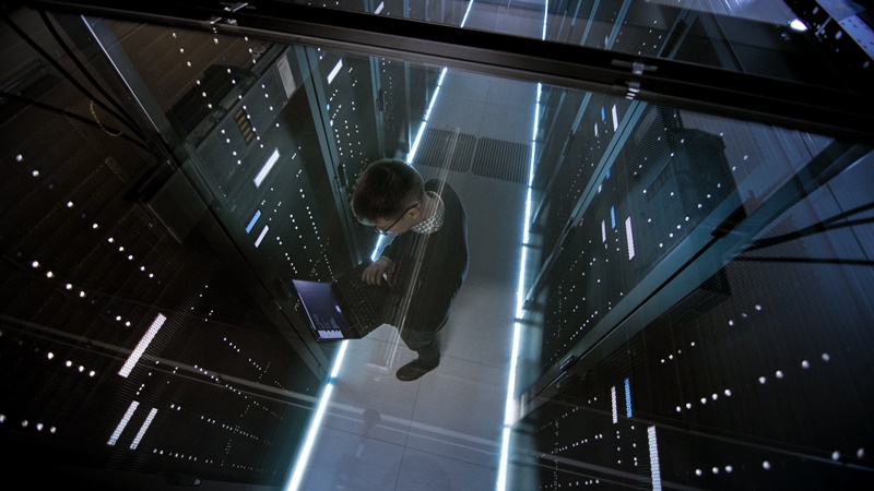 Top View Through the Glass of IT Engineer Working with Laptop in Data Center Full of Active Rack Servers.